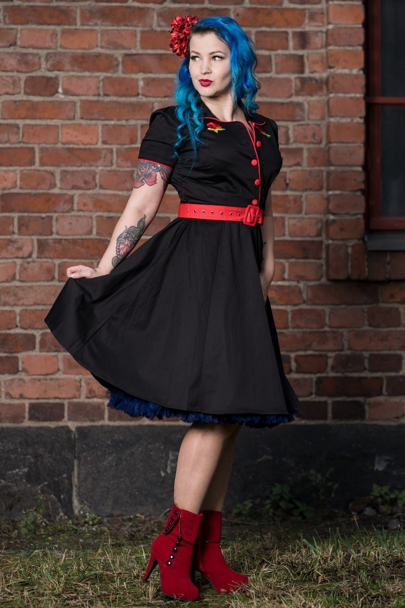 Blue haired woman wears our short sleeved Sherry diner dress in black, with red buttons, belt and roses, in front of a brick wall