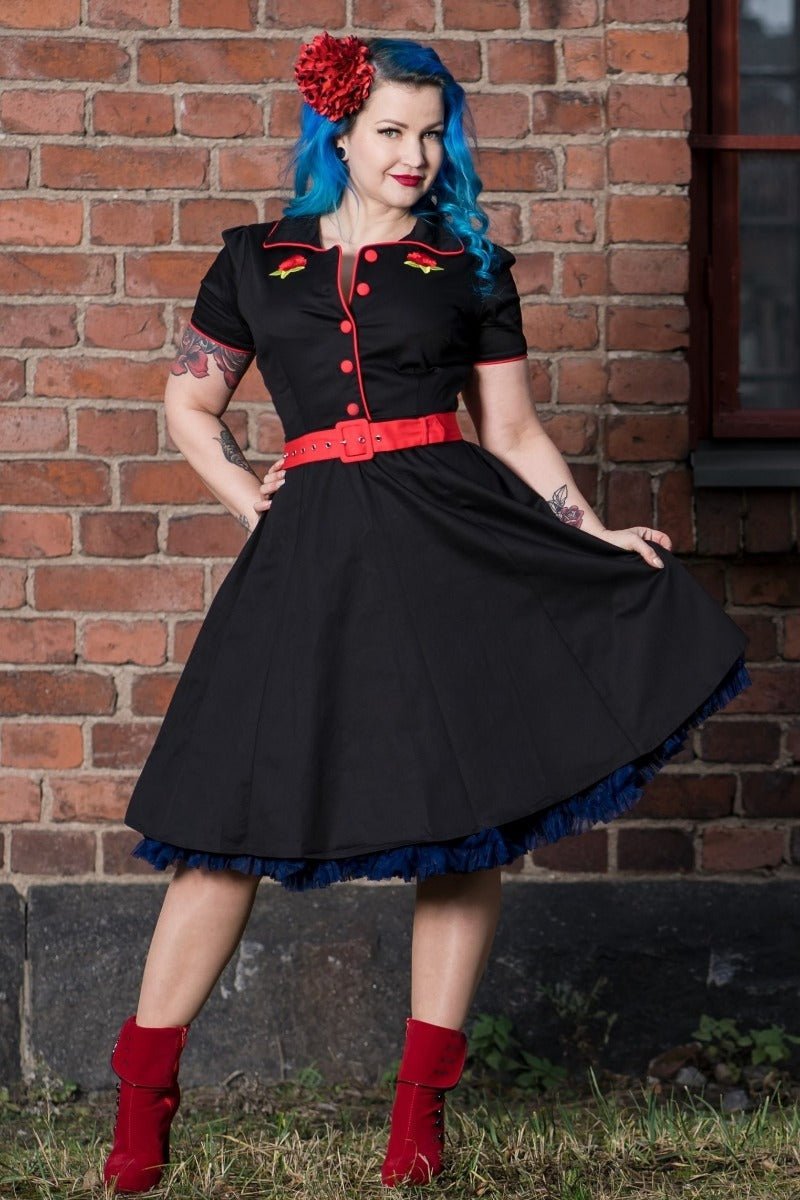 Woman wears our short sleeved Sherry diner dress in black, with red buttons, belt and roses, in front of a brick wall